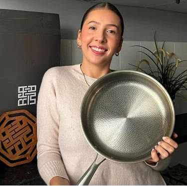Smiling woman holding Siraat Titanium Hammered Pan Pro in her kitchen, showing the hand-hammered non-stick surface