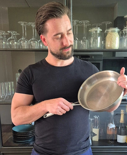 Man admiring the hammered titanium surface of his Siraat pan in a modern kitchen with glassware shelving